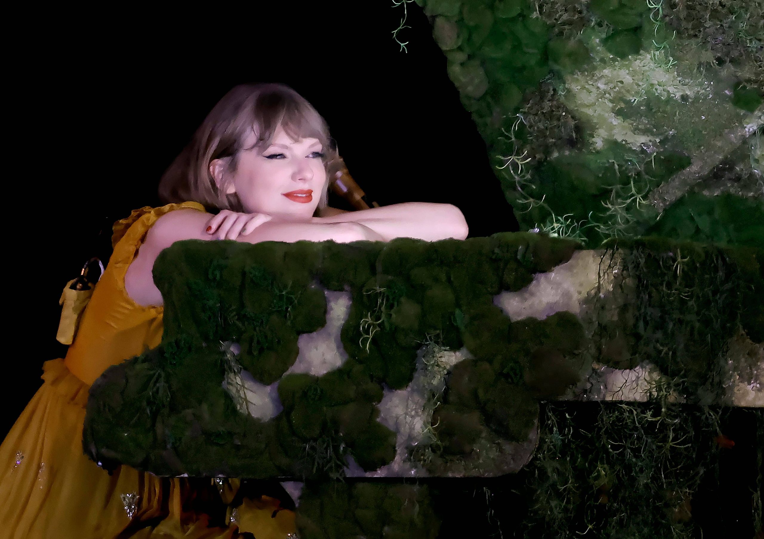 Taylor Swift in yellow dress leans on a moss-covered piano, smiling serenely. Dark background enhances the lush green texture. 