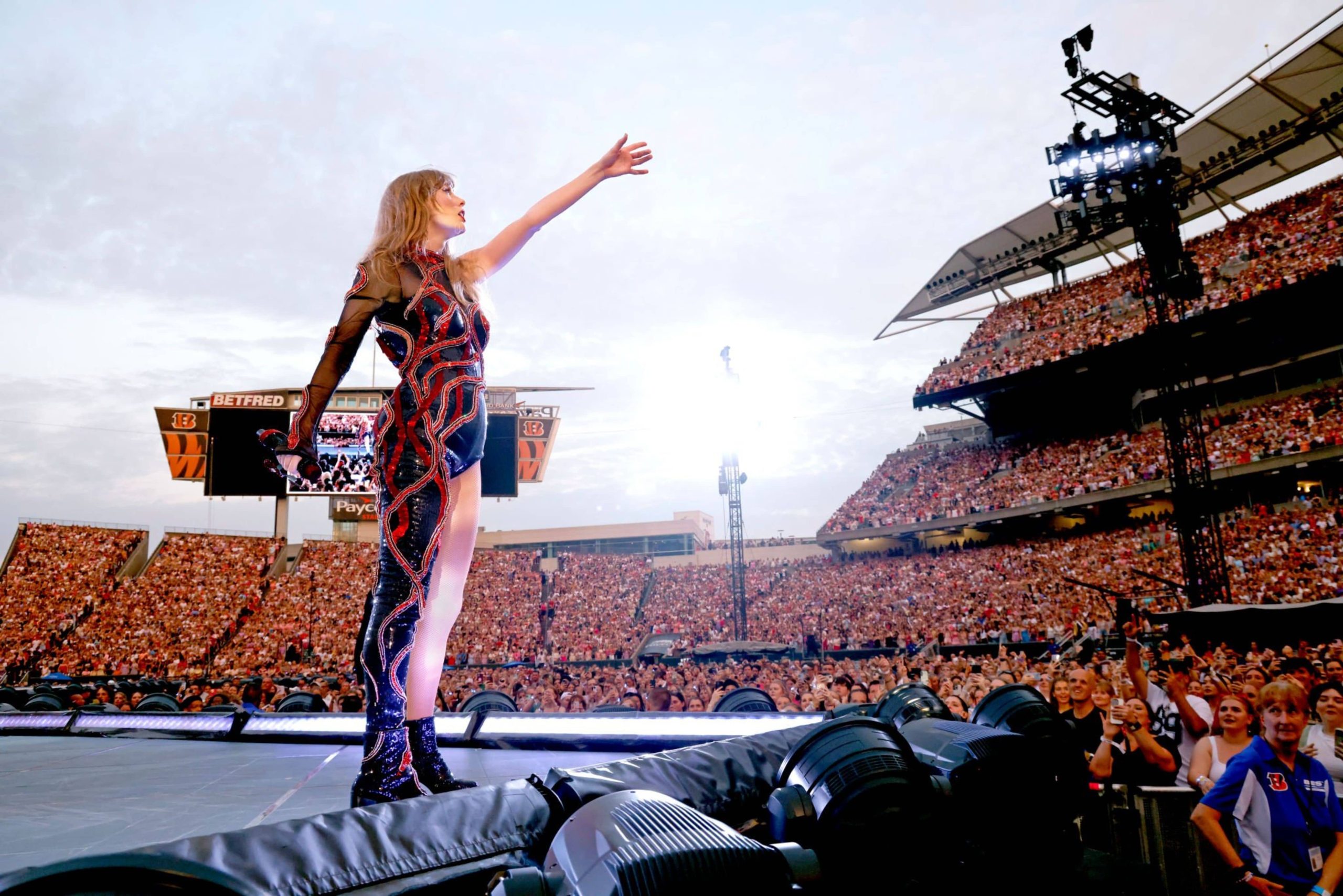 Taylor Swift in a black and red outfit performs on stage in a packed stadium, arm outstretched, with a bright sky and enthusiastic crowd.