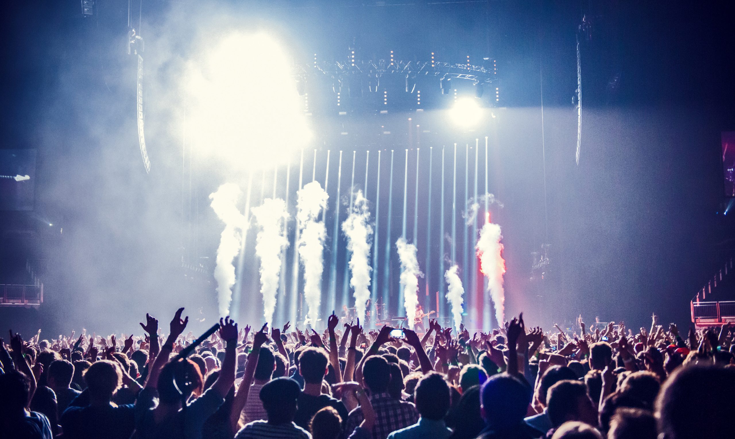 Concert crowd with hands raised, vibrant stage lights and smoke effects. Energetic atmosphere with blue and red hues in a dark arena.