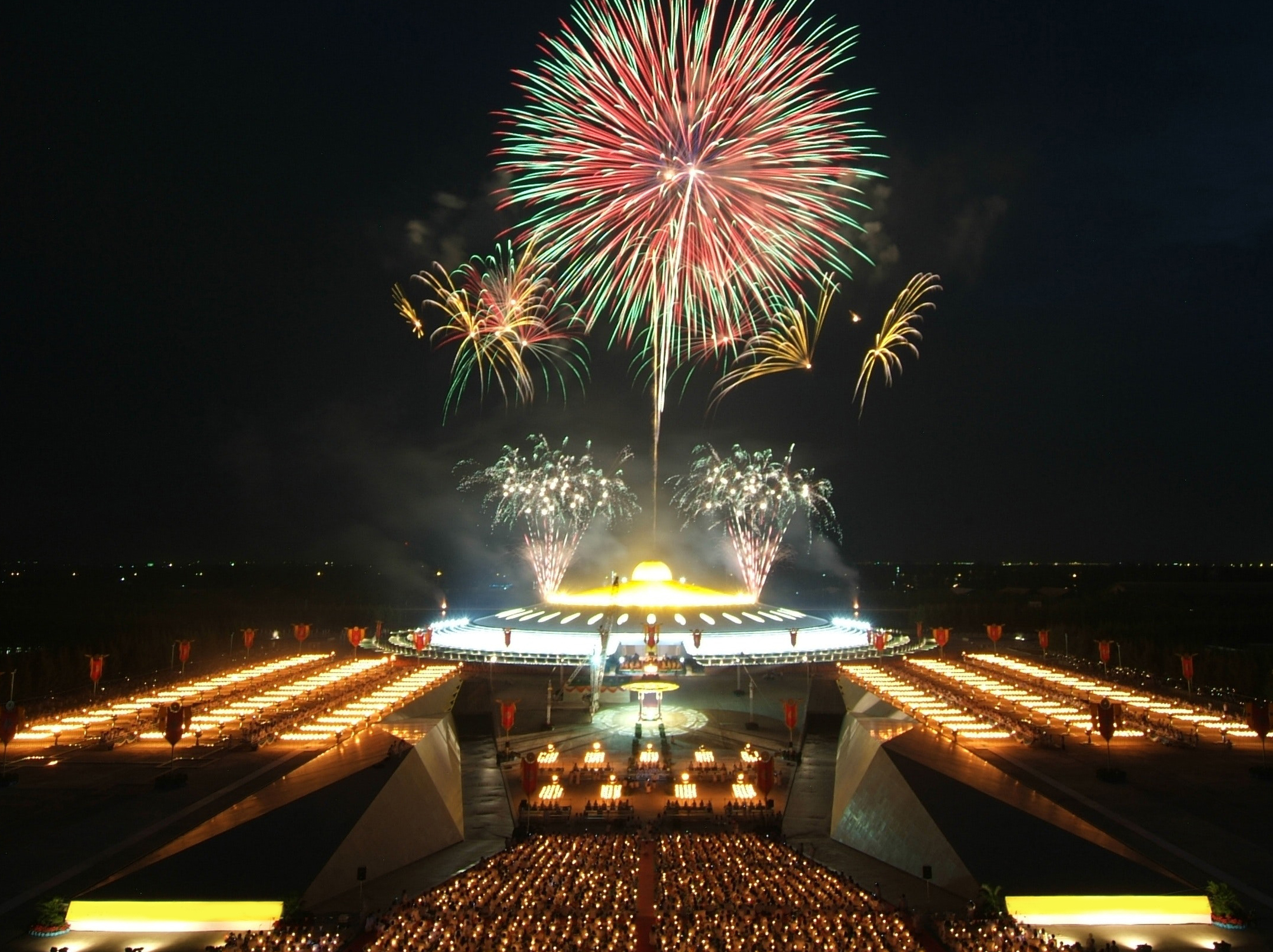 Fireworks burst in colorful patterns over a large, illuminated circular structure. Rows of lights and a crowd create a festive atmosphere.
