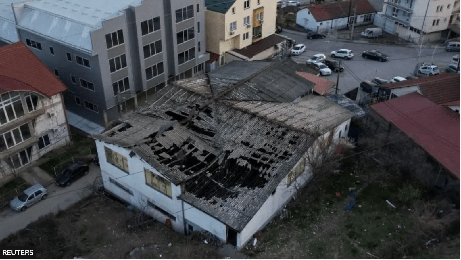 Decaying building with a large, hole-filled roof in an urban area. Surrounding buildings show residential and industrial architecture. Overcast mood.