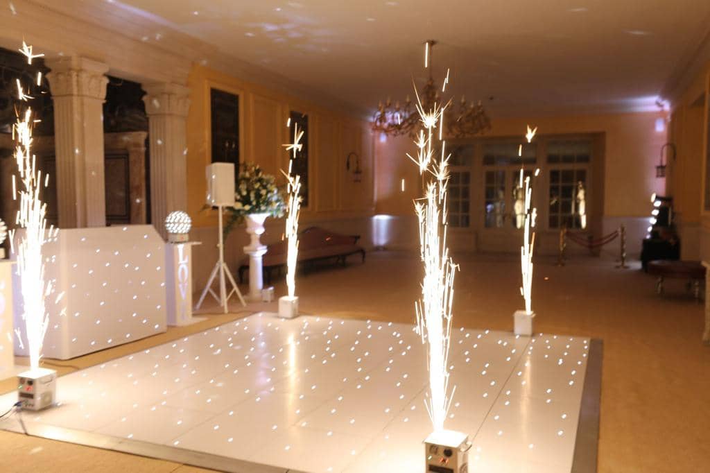 Indoor event space with sparklers on a lit floor. Columns and chandeliers in background. A sign with "LOVE" and white flowers are present.