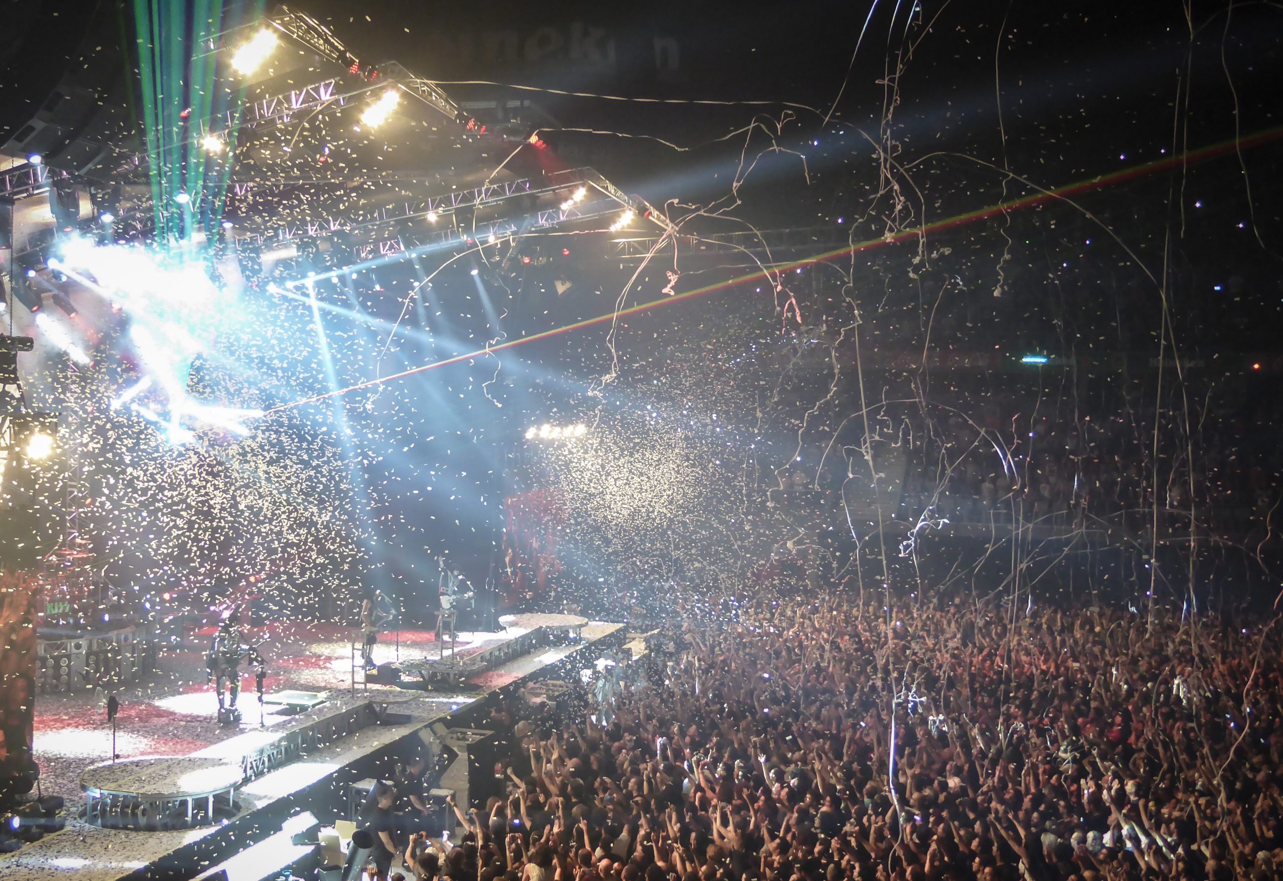 Concert scene with a band on stage under bright lights, confetti falling, and a large crowd cheering. Energetic and festive atmosphere.