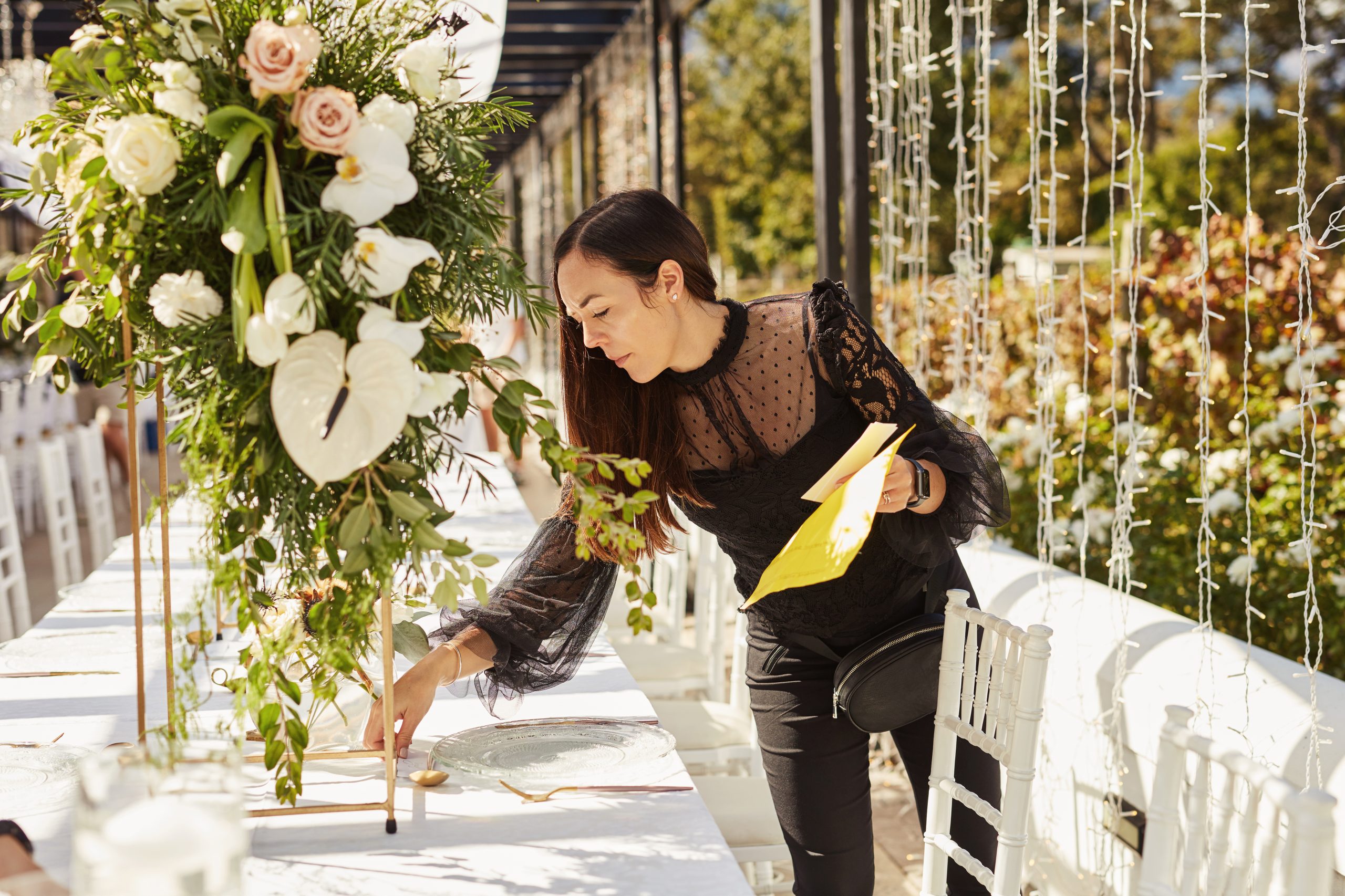 Woman in black dress sets table with floral centerpiece outside under string lights, holding yellow paper, creating an elegant setup.