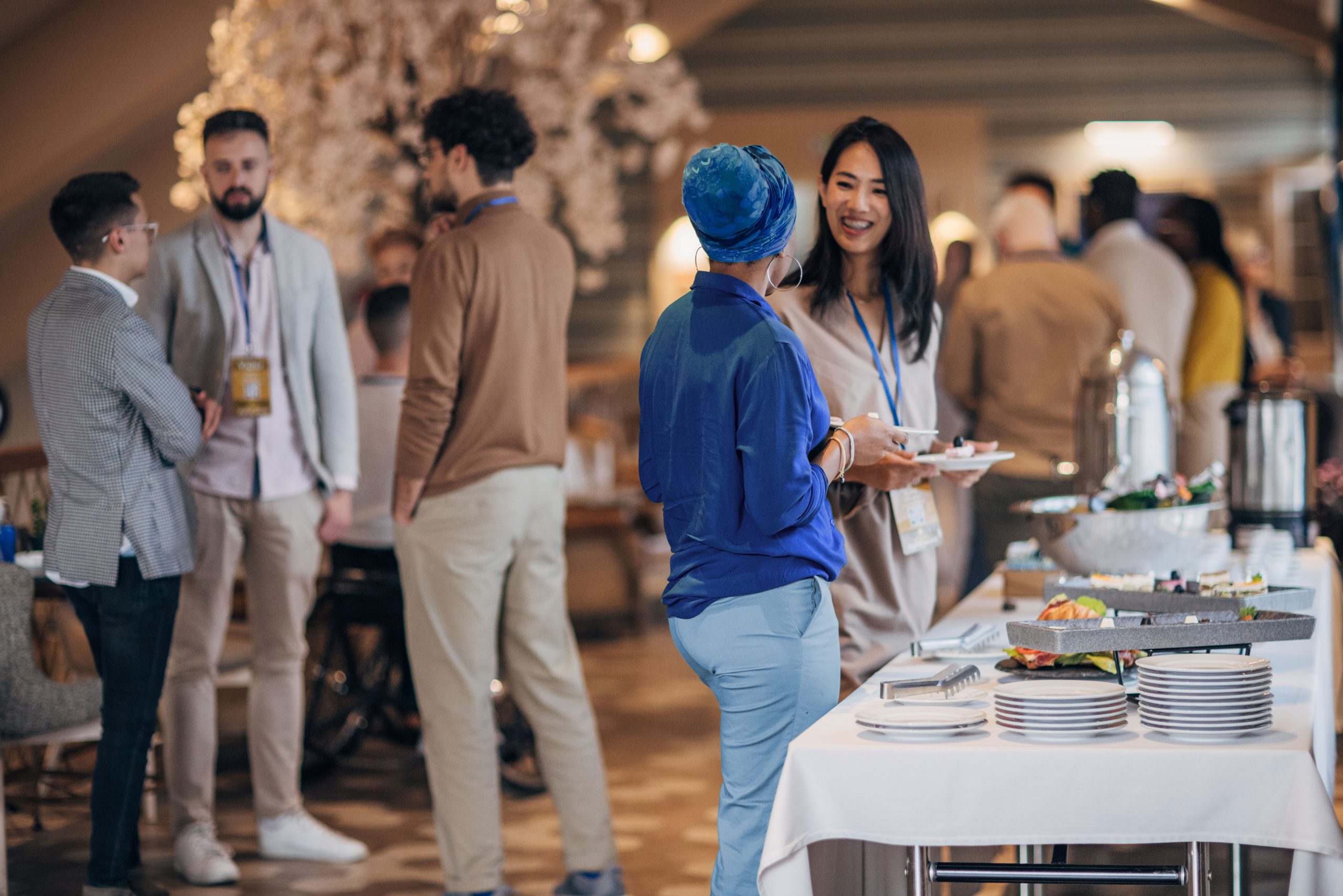People mingling at a conference buffet; two women smiling in foreground, others chatting. Bright lighting, elegant setting, name tags visible.