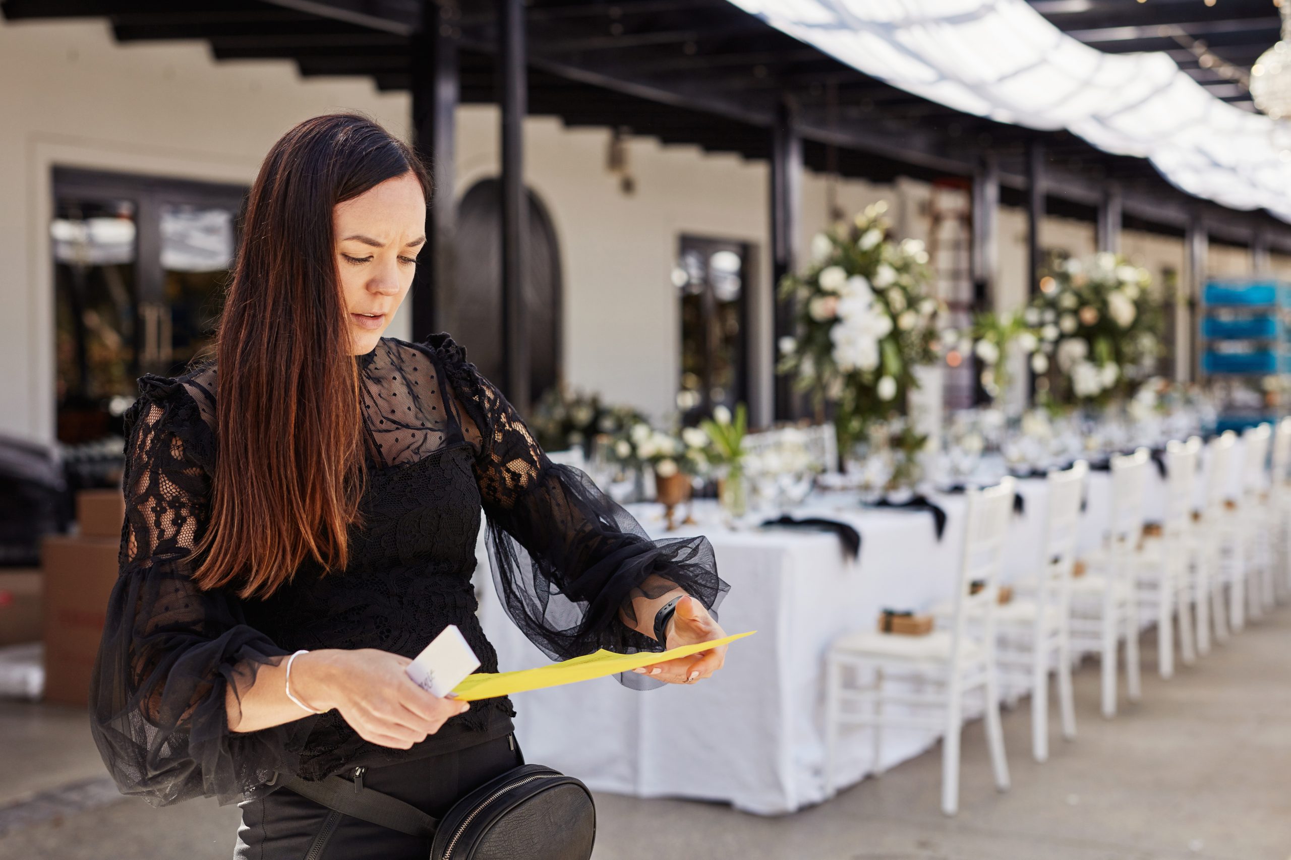 Woman in black lace top holds yellow paper, standing in a decorated outdoor venue with white tables and floral arrangements. Focused expression.