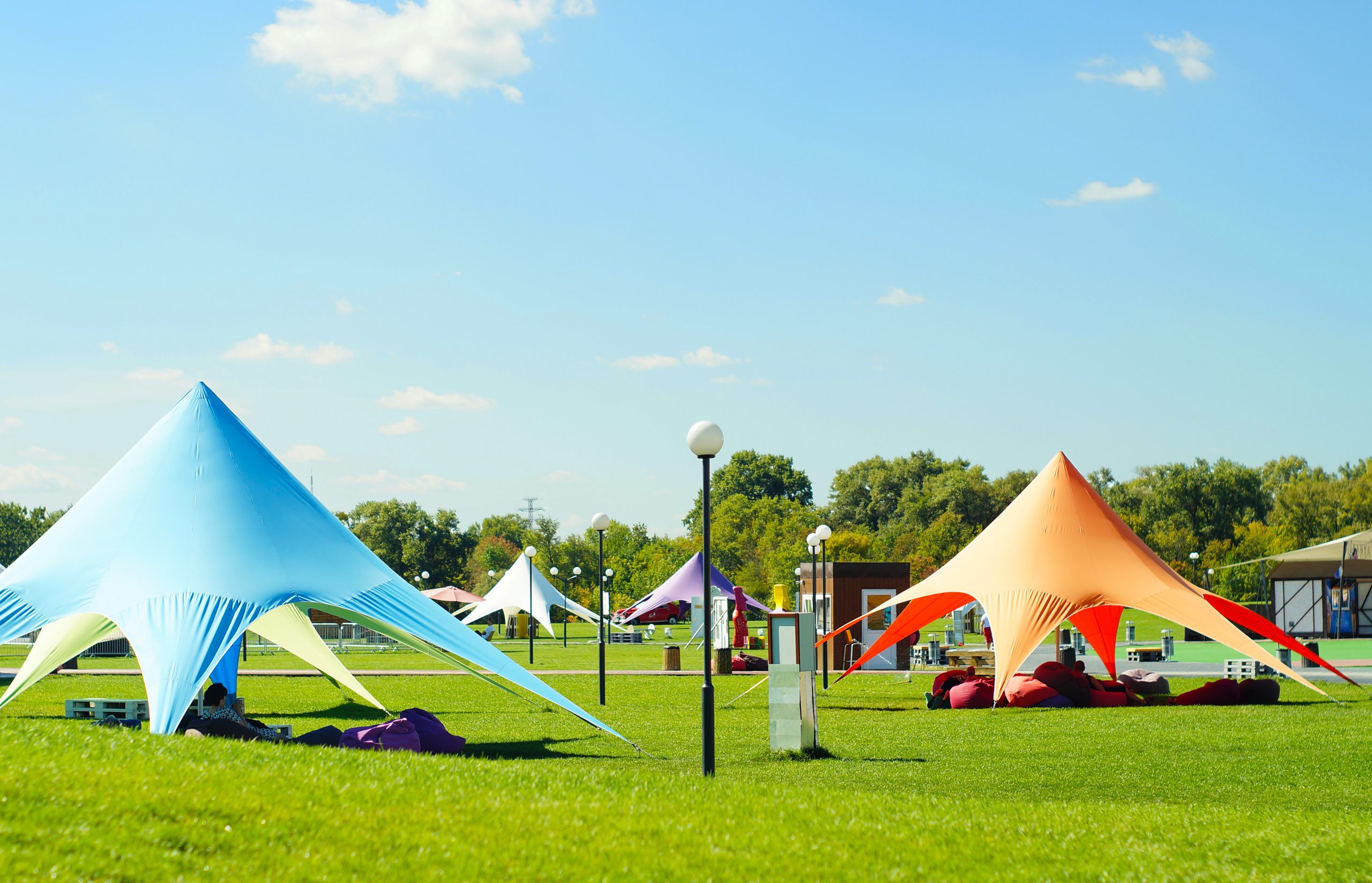 Colorful tents in a sunny park with green grass and blue sky. People relax on bean bags under blue and orange canopies.