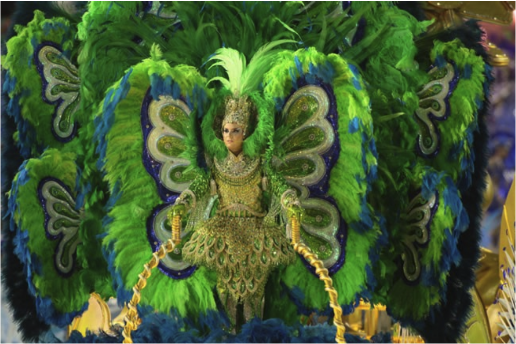 Person in elaborate green and blue costume with feathered wings and intricate patterns, set against a vibrant festival backdrop.