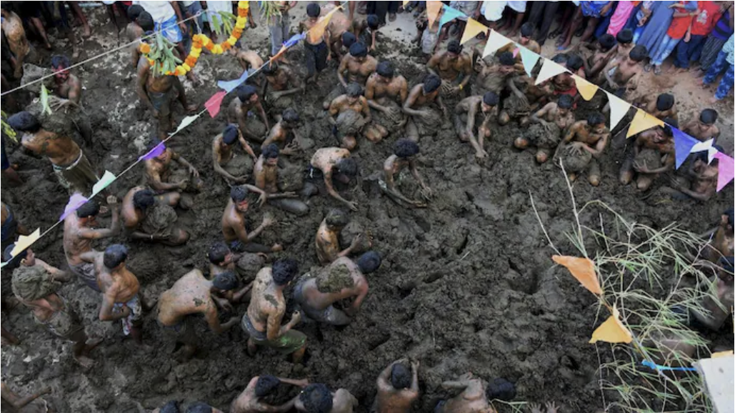 Men covered in mud participate in a wrestling match in a muddy pit, surrounded by colorful flags and spectators, creating a festive vibe.
