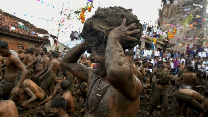 People covered in mud participate in a festival, smiling and holding mud balls. Colorful flags and crowds in the background create a lively scene.