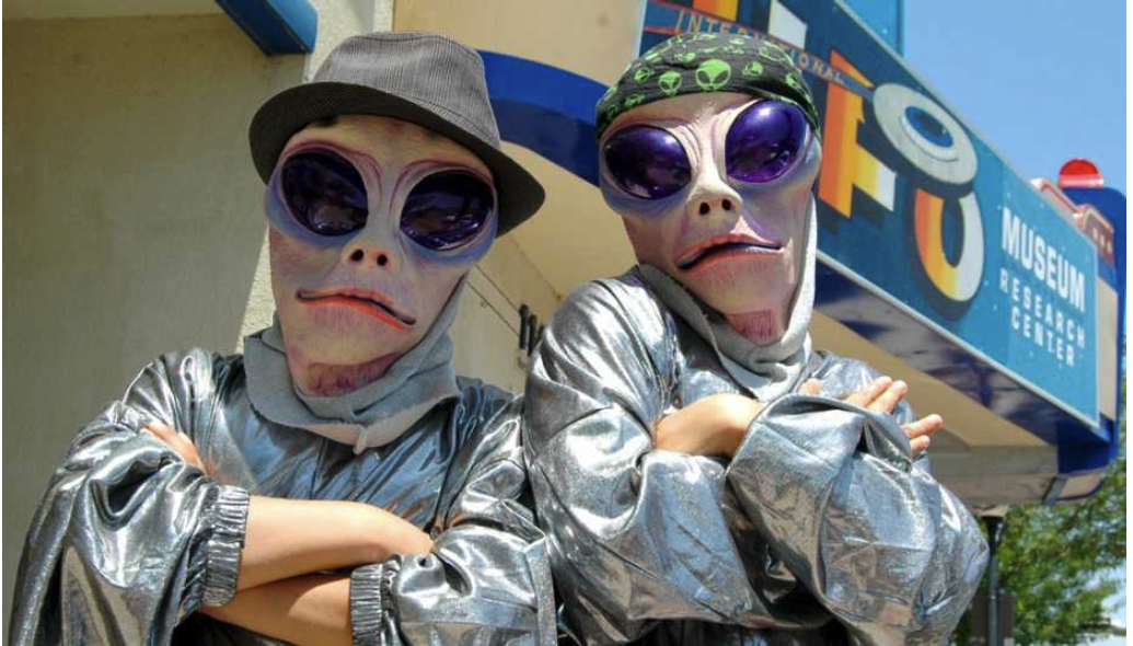 Two people in alien costumes with large sunglasses, silver outfits, and hats pose in front of a UFO Museum sign, a sunny day.