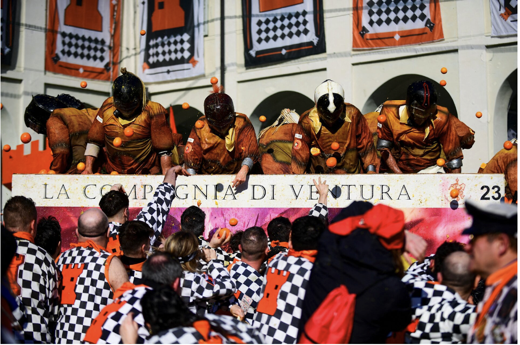 People in colorful outfits throw oranges in a lively street festival. A sign reads "LA COMPAGNIA DI VENTURA." Vibrant, chaotic scene.