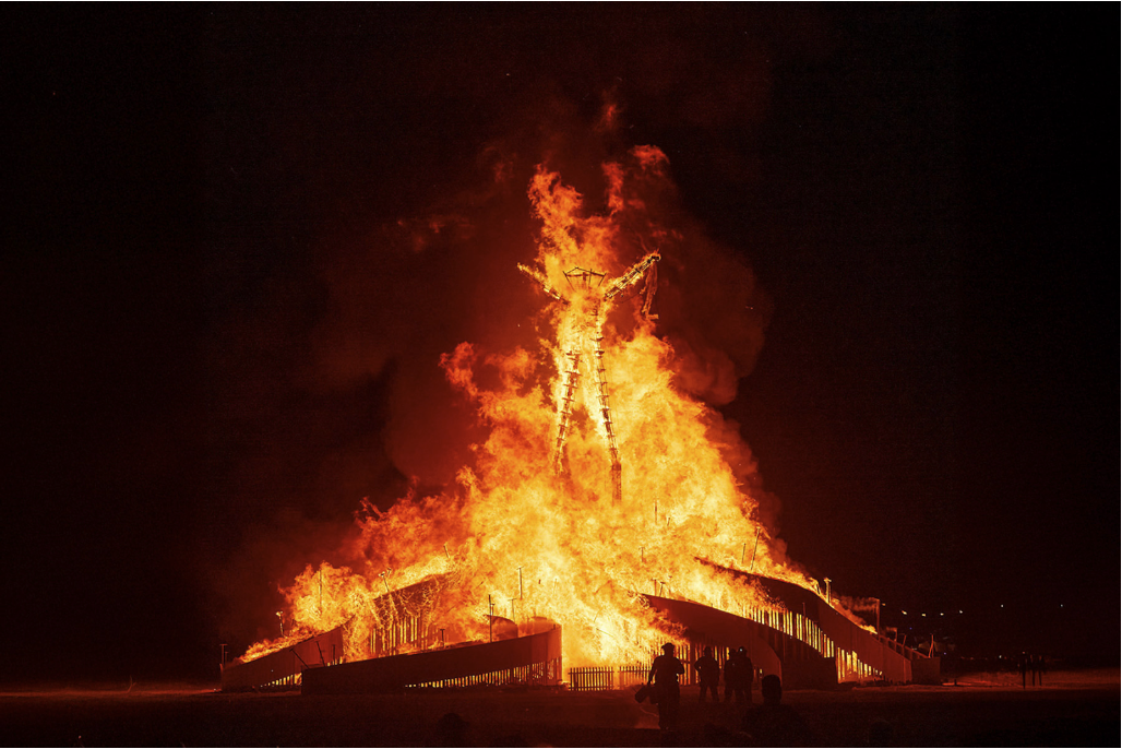 A large wooden figure burns brightly at night, surrounded by silhouetted spectators. Intense orange flames illuminate the dark sky.