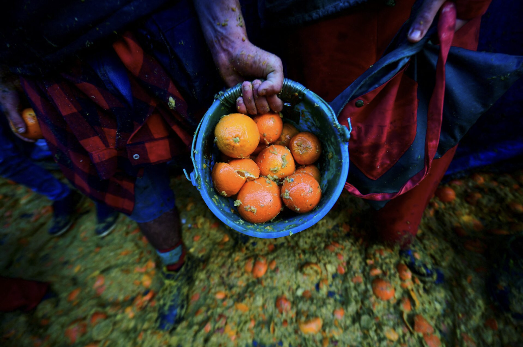 A person holds a blue bucket of oranges, some broken, during a festival or event. Ground and clothing are splattered with orange pulp.