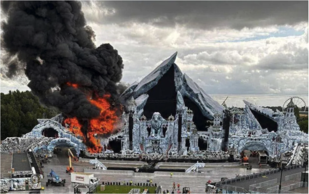 Stage facade on fire with large flames and black smoke against a cloudy sky. Ice-themed design, ferris wheel visible in the background.