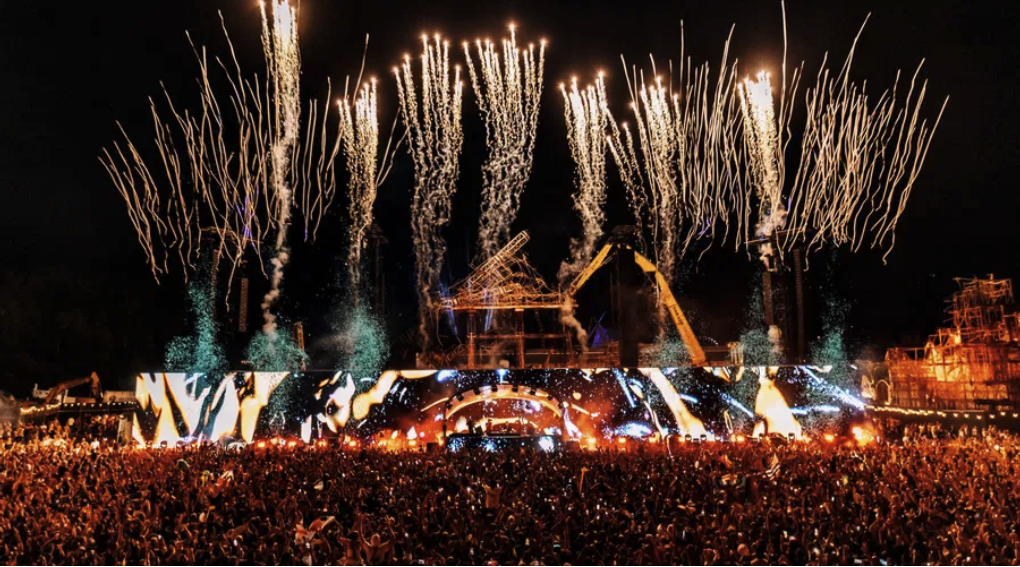 Crowd at a nighttime music festival with colorful stage lights and fireworks. The mood is energetic and festive.