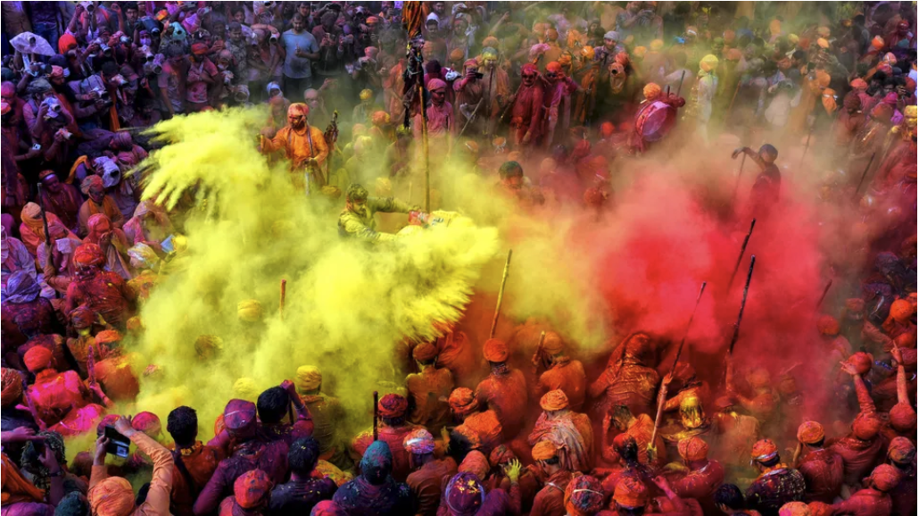 A vibrant crowd celebrates Holi, throwing yellow and red powder. People in colorful attire, some holding sticks, with joyful energy.
