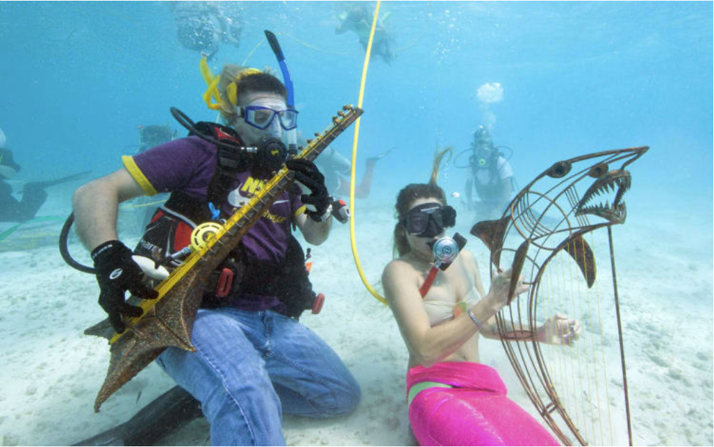 Divers underwater playing fish-shaped instruments; one wears purple, the other pink. Clear ocean setting, others in background, joyful vibe.