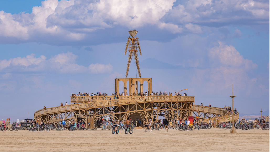 Crowd gathers at a large wooden sculpture under a cloudy blue sky. Bicycles and people surround the structure, creating a festive atmosphere.