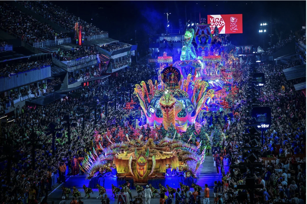 Colorful parade with vibrant floats, performers, and crowd. Bright lights at night create a festive, lively atmosphere. Visible text: "VIRA DOU."
