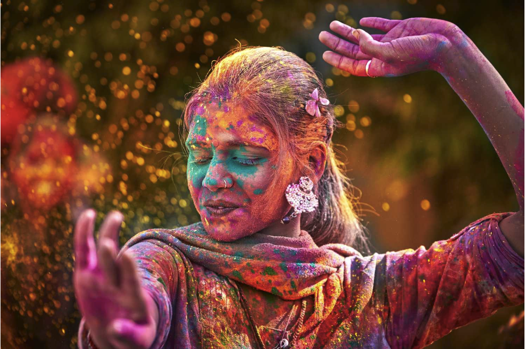 A joyful woman with closed eyes is covered in vibrant colored powders during a Holi celebration, set against a blurred background.